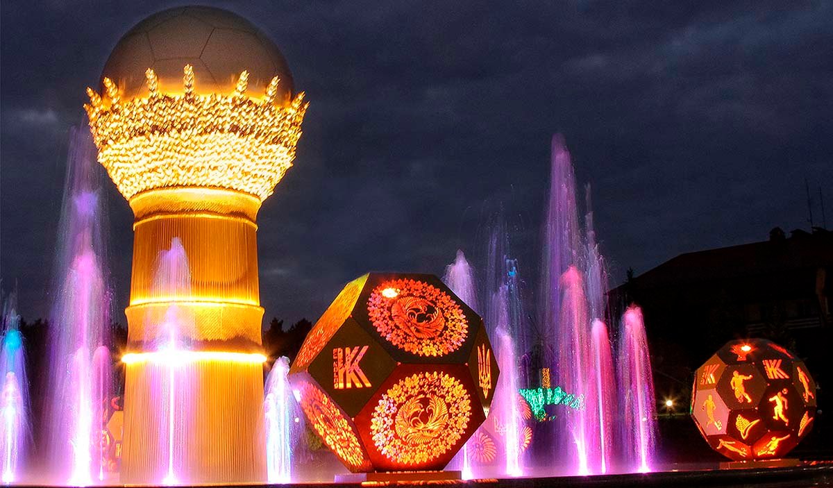 Light-dynamic public art with fountains, a wheat sheaf sculpture, and illuminated geometric sculptures in the town square at night