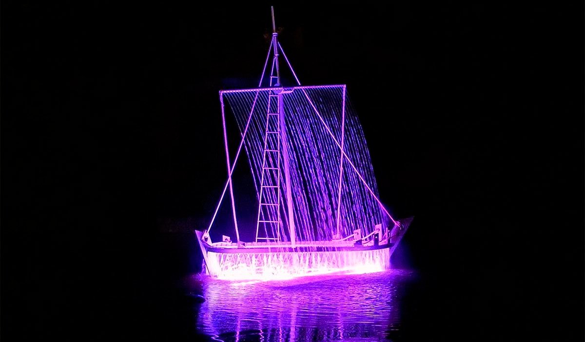 A dynamic public art piece in the shape of a sailing ship illuminated in purple and pink on the water at night.