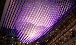 A kinetic installation of hundreds of teardrop-shaped units illuminated in purple, forming a three-dimensional face beneath the glass dome of a modern atrium.