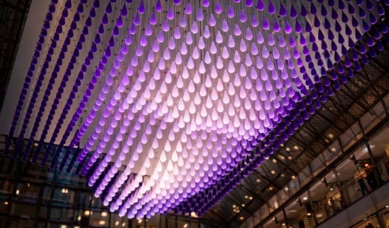 A kinetic installation of hundreds of teardrop-shaped units illuminated in purple, forming a three-dimensional face beneath the glass dome of a modern atrium.