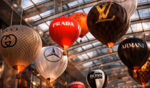 Кинетическая инсталляция «Shopping Vortex» — подвесная композиция из брендированных воздушных шаров Prada, Louis Vuitton, Gucci, Armani, Boss и Mercedes под стеклянным куполом, с тёплой подсветкой, создающей эффект роскошного вихря покупок.