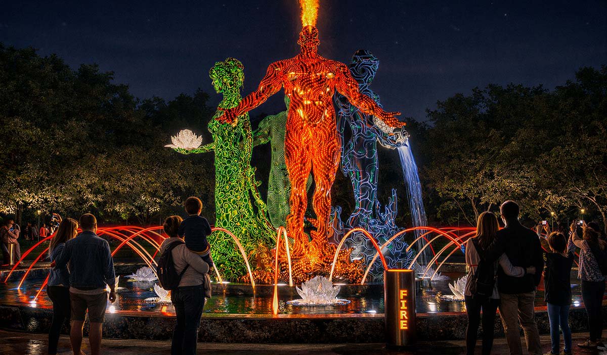 A public art fountain with sculptures of people of different colors is surrounded by people in the park in the evening.